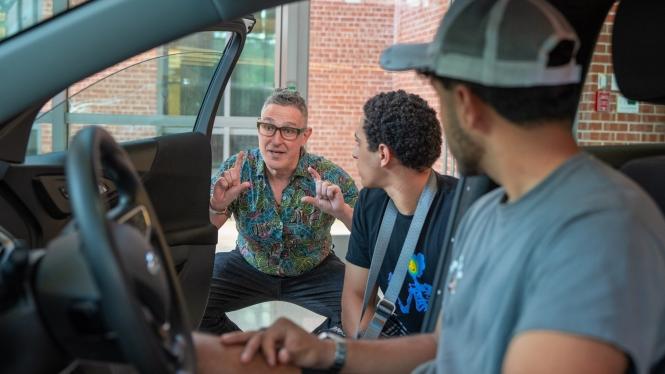 Professor leans in front of open vehicle door with two students inside car